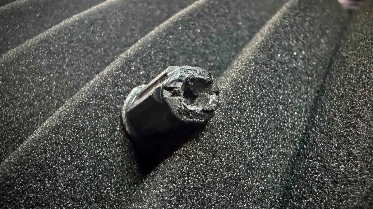 A black Rackstuds lock nut with its head torn and chewed open, stripped by an impact driver, sitting on grey acoustic foam